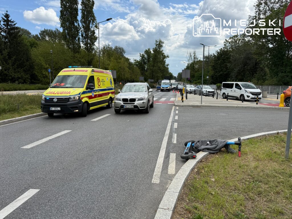 Ambulans zatrzymany przy drodze, obok leżąca skutera, a w tle przejeżdżające samochody.