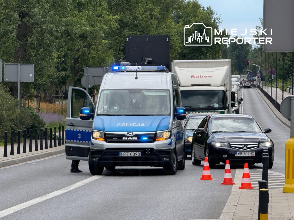 Policyjny radiowóz z otwartymi drzwiami kontroluje ruch drogowy, a w tle widoczne są samochody oraz sygnalizacja świetlna.
