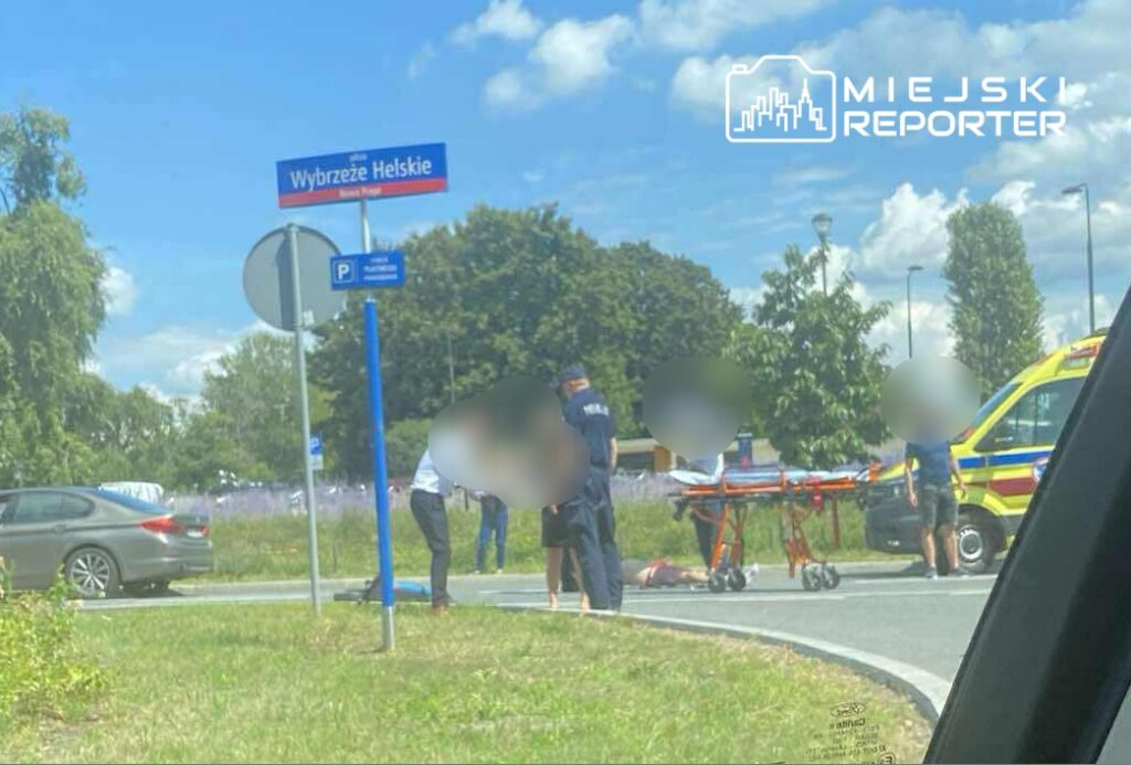 Grupa ratowników medycznych udziela pomocy osobie leżącej na drodze obok karetki na skrzyżowaniu.