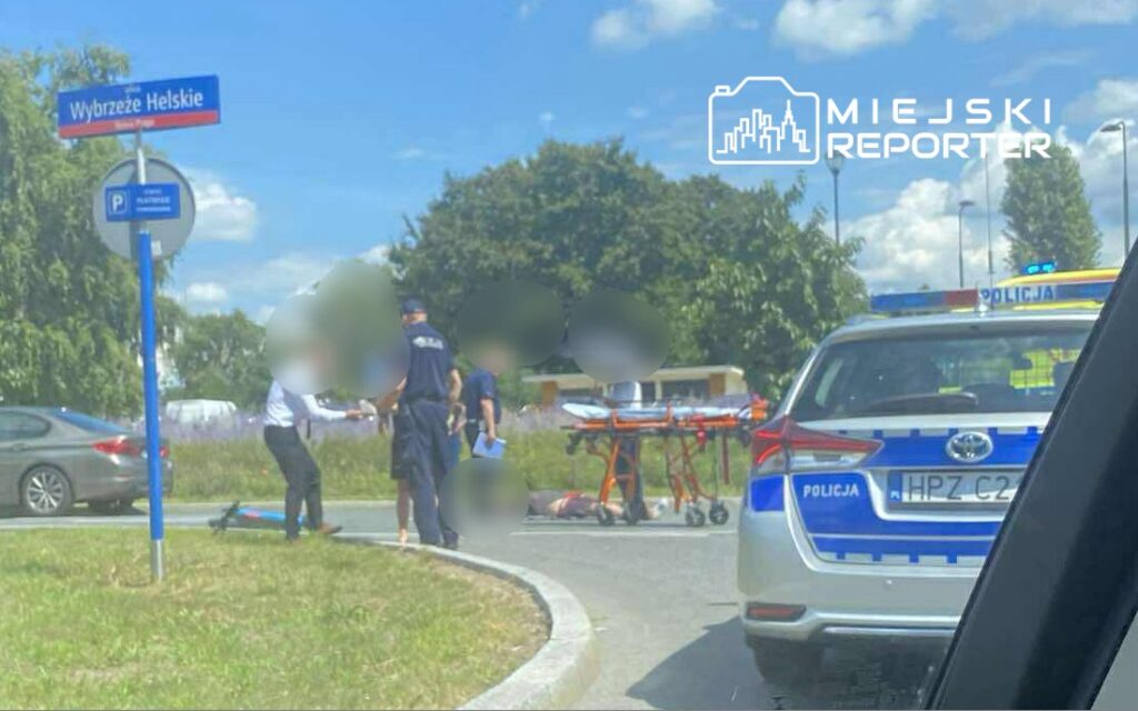 Mężczyzna w garniturze rozmawia z policjantem obok leżącego na ziemi mężczyzny, w tle widać radiowóz.
