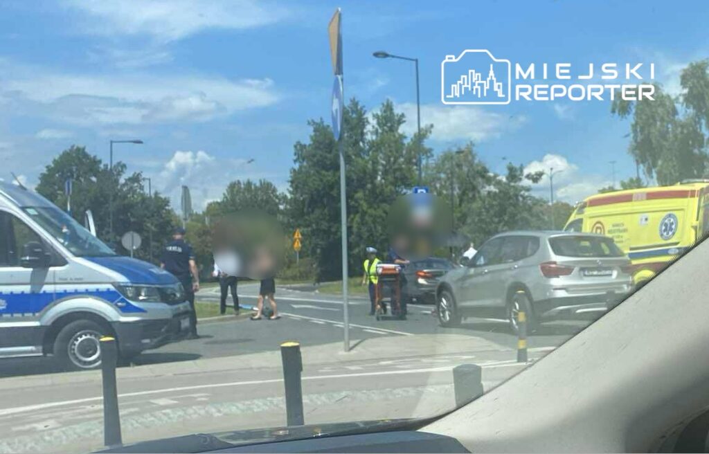 Na rondzie kilka osób otacza rannego mężczyznę, obok stoi policyjny radiowóz i karetka.
