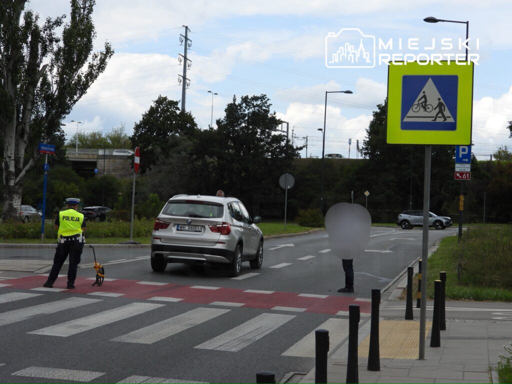 Policjant mierzy odległość na przejściu dla pieszych, obok zatrzymany samochód osobowy. W tle widać znaki drogowe i zieleń.