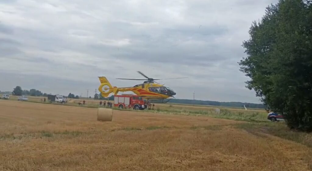 Helikopter ratunkowy ląduje na polu z zebranym zbożem obok wozu strażackiego i grupy osób.