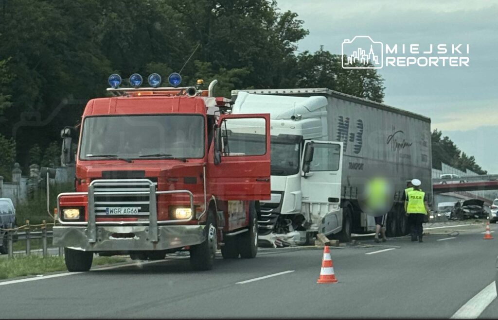 Czerwony wóz strażacki stoi obok zderzonej ciężarówki na drodze, w otoczeniu oznaczeń policyjnych i pachołków.