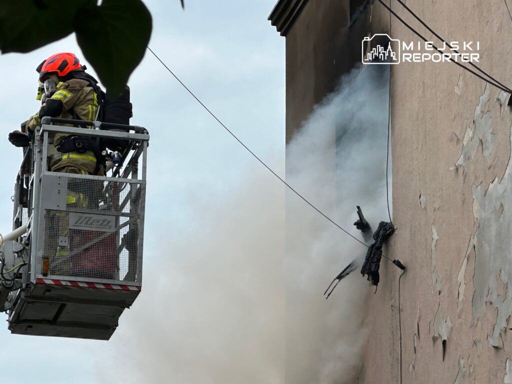 Strażacy w podnośniku gaszą pożar na ścianie budynku, z którego wydobywa się dym.