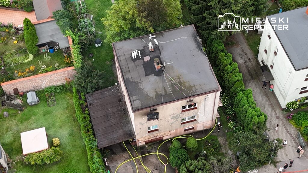 Dwupiętrowy budynek z uszkodzoną elewacją, otoczony zielenią, z wężami strażackimi na ziemi i ludźmi w pobliżu.