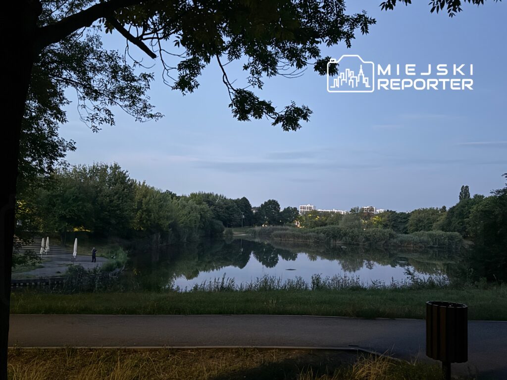 Mężczyzna stojący na brzegu stawu, otoczony zielenią, w tle odbicie drzew na wodzie.