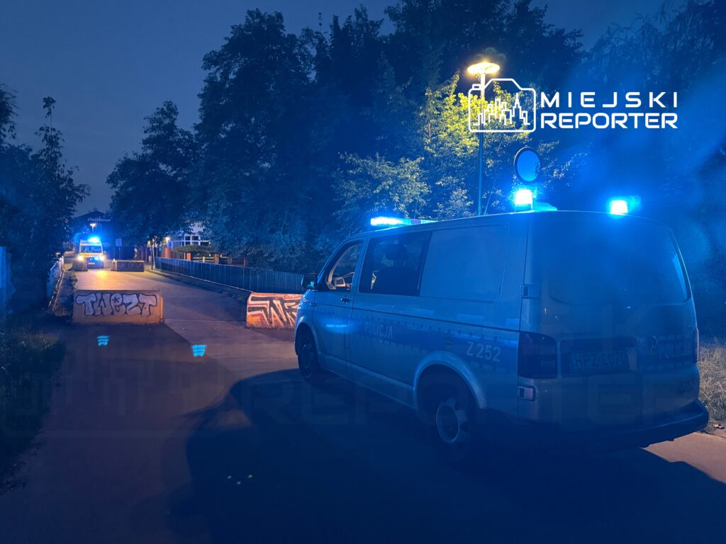 Policyjny van z niebieskimi światłami zatrzymany przy barierkach z graffiti na drodze w nocy.