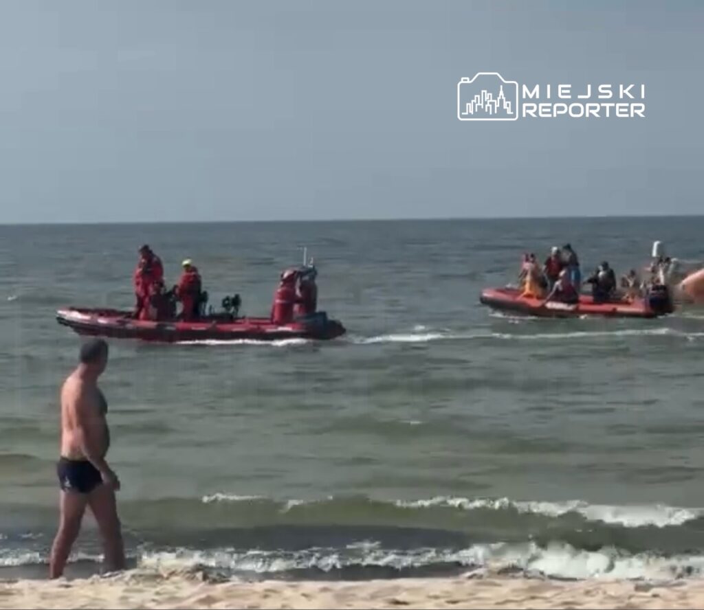 Mężczyzna w kąpielówkach spacerujący po plaży, obok przepływające łodzie ratunkowe z ratownikami.