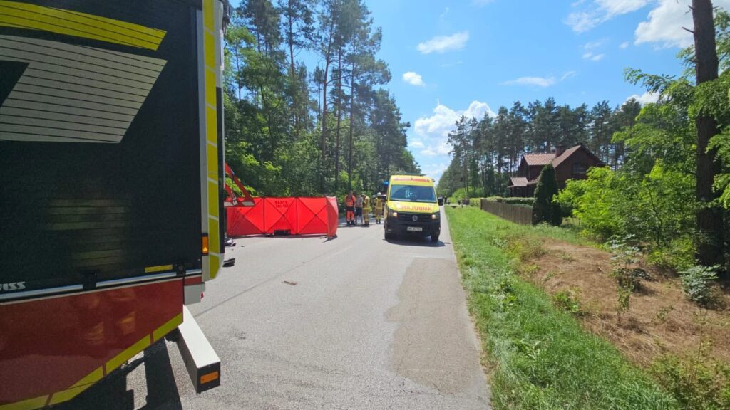 Zespół ratunkowy pracuje przy drodze, otoczony przez czerwoną barierkę, w otoczeniu lasu i domów.