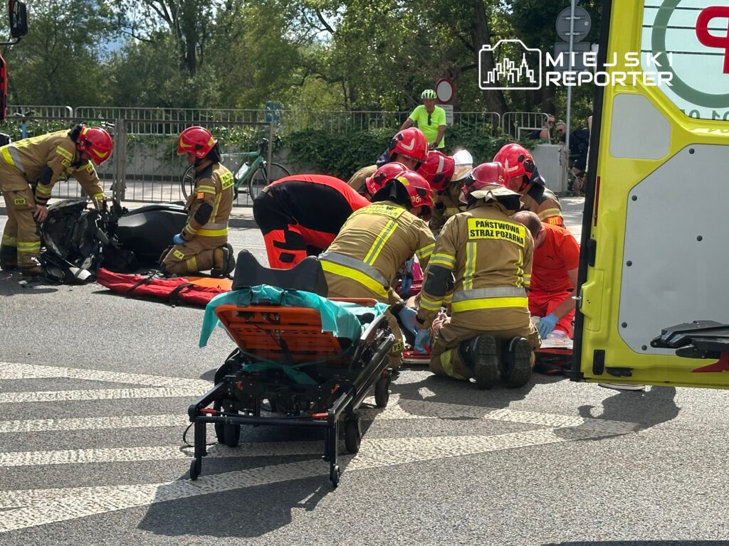 Grupa ratowników w mundurach pracuje nad poszkodowanym leżącym na noszach na ulicy, obok przewrócony motocykl.