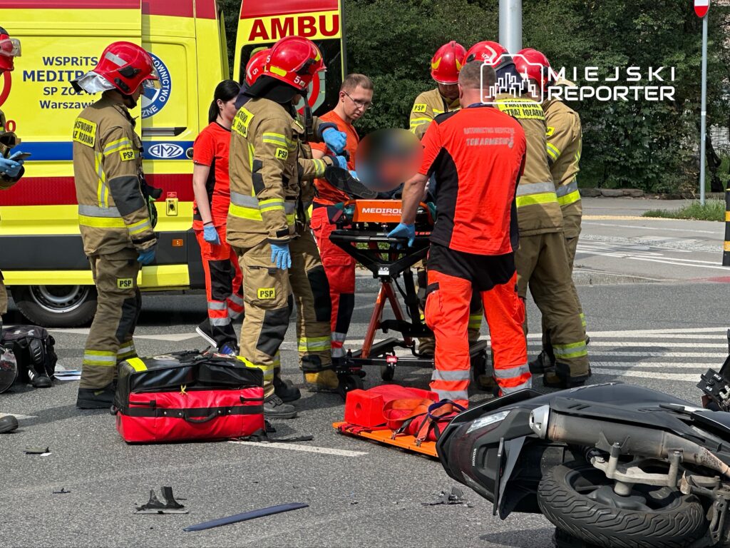 Grupa ratowników medycznych udziela pomocy poszkodowanemu na jezdni obok przewróconego skutera.