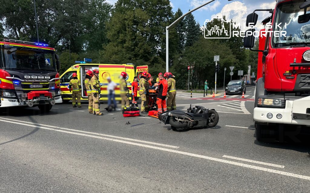 Grupa ratowników medycznych i strażaków udziela pomocy poszkodowanemu obok przewróconego motocykla na skrzyżowaniu.