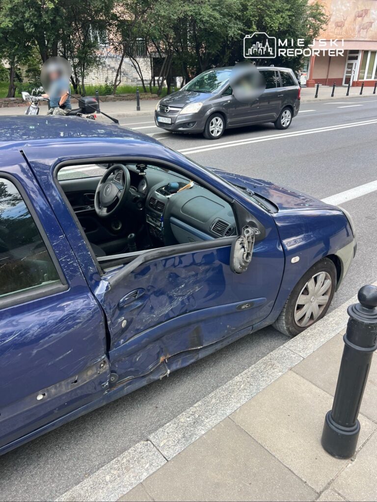 Uszkodzony niebieski samochód osobowy stoi na ulicy, z otwartymi drzwiami i widoczną dentą na lewym boku.