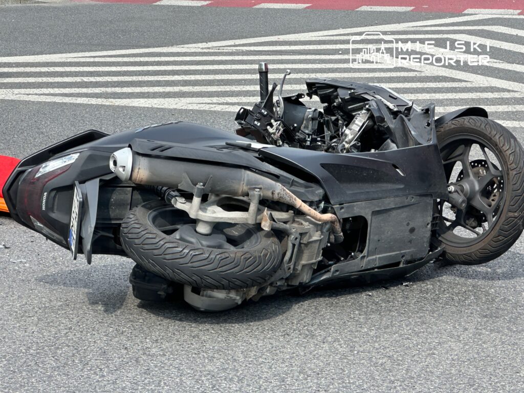 Motocykl leżący na boku na jezdni, z uszkodzoną konstrukcją i wygiętą kierownicą. W tle widoczne pasy drogowe.