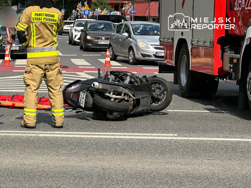Strażak w mundurze stoi obok przewróconego motocykla na ruchliwej ulicy, w tle widoczne samochody i służby ratunkowe.