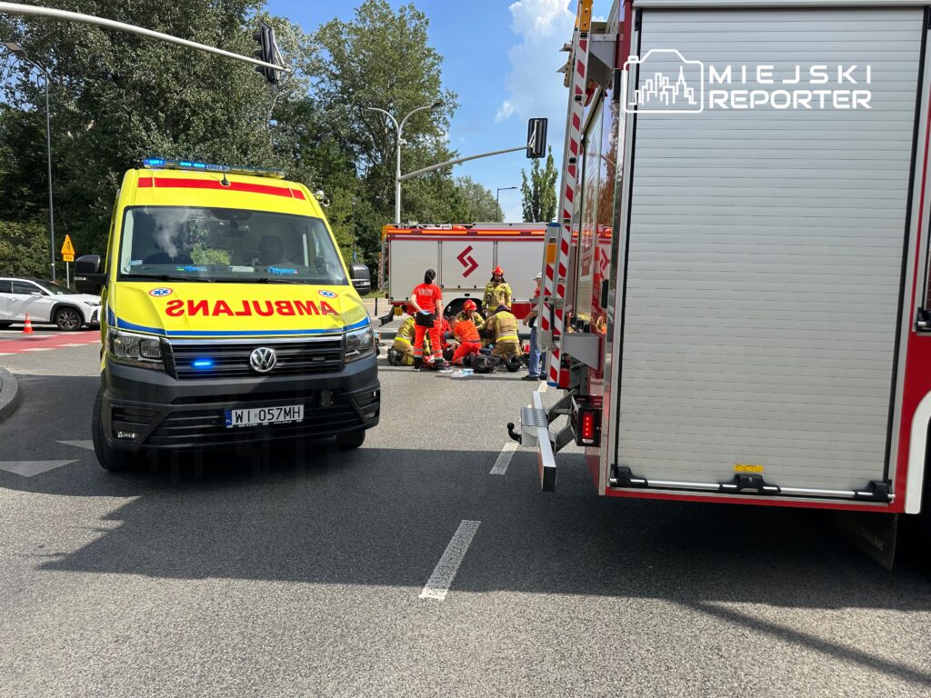 Karetka z włączonymi sygnałami świetlnymi zatrzymana obok grupy ratowników udzielających pomocy poszkodowanemu na drodze.
