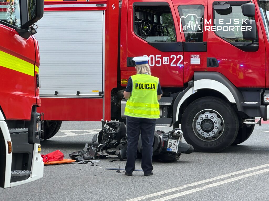 Policjantka w kamizelce odblaskowej bada wypadek drogowy obok wozu strażackiego, na jezdni leżą fragmenty motocykla.