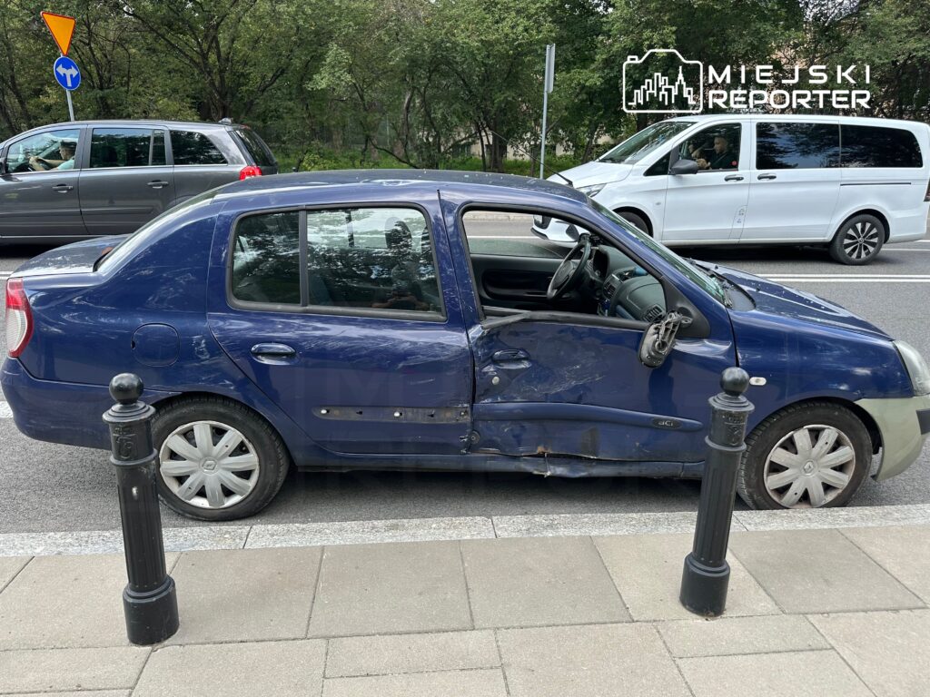 Zniszczony niebieski samochód osobowy zaparkowany na chodniku, z otwartymi drzwiami i uszkodzoną boczną częścią.