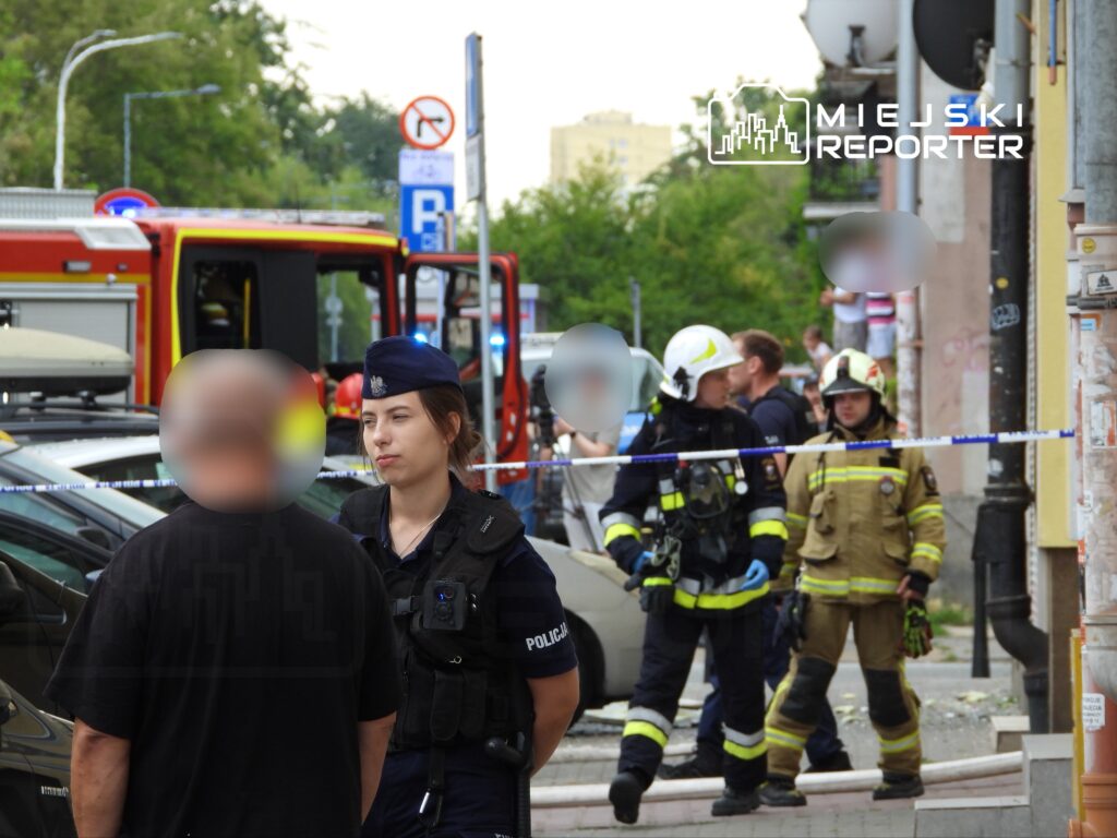 Policjantka rozmawia z mężczyzną na ulicy, w tle strażacy i woz strażacki, miejsce zabezpieczone taśmą.