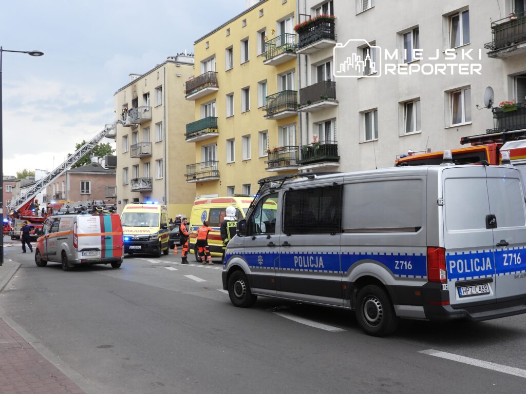 Na ulicy z widocznymi pojazdami straży pożarnej i ambulansami, ratownicy interweniują przy budynku mieszkalnym.