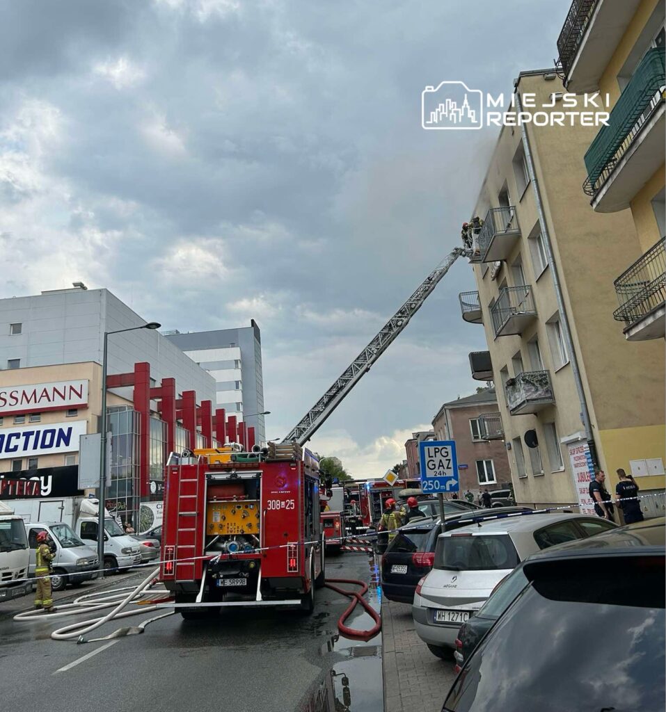 Strażacy na drabinie gaszą pożar na balkonie w budynku mieszkalnym, obok stoi wóz strażacki.
