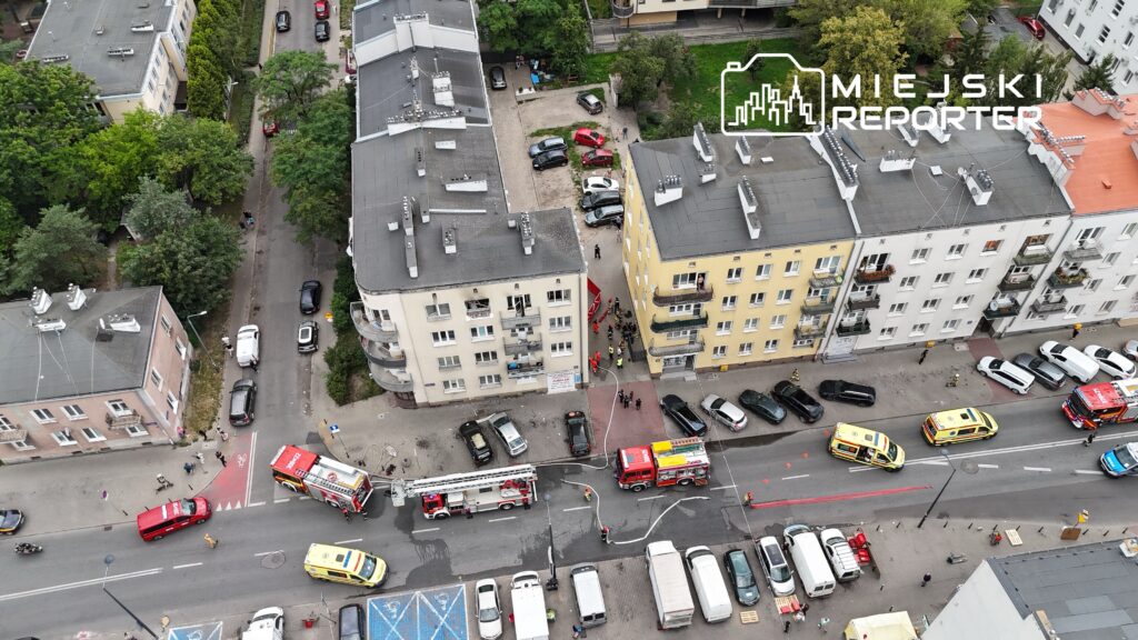 Wielka akcja ratunkowa w miejskiej okolicy; strażacy i ratownicy medyczni pracują przy wozach strażackich na ulicy.