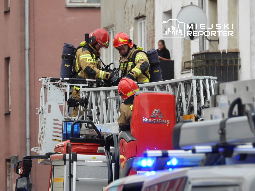 Strażacy w hełmach pracują na podnośniku strażackim, przygotowując sprzęt w pobliżu budynku mieszkalnego.