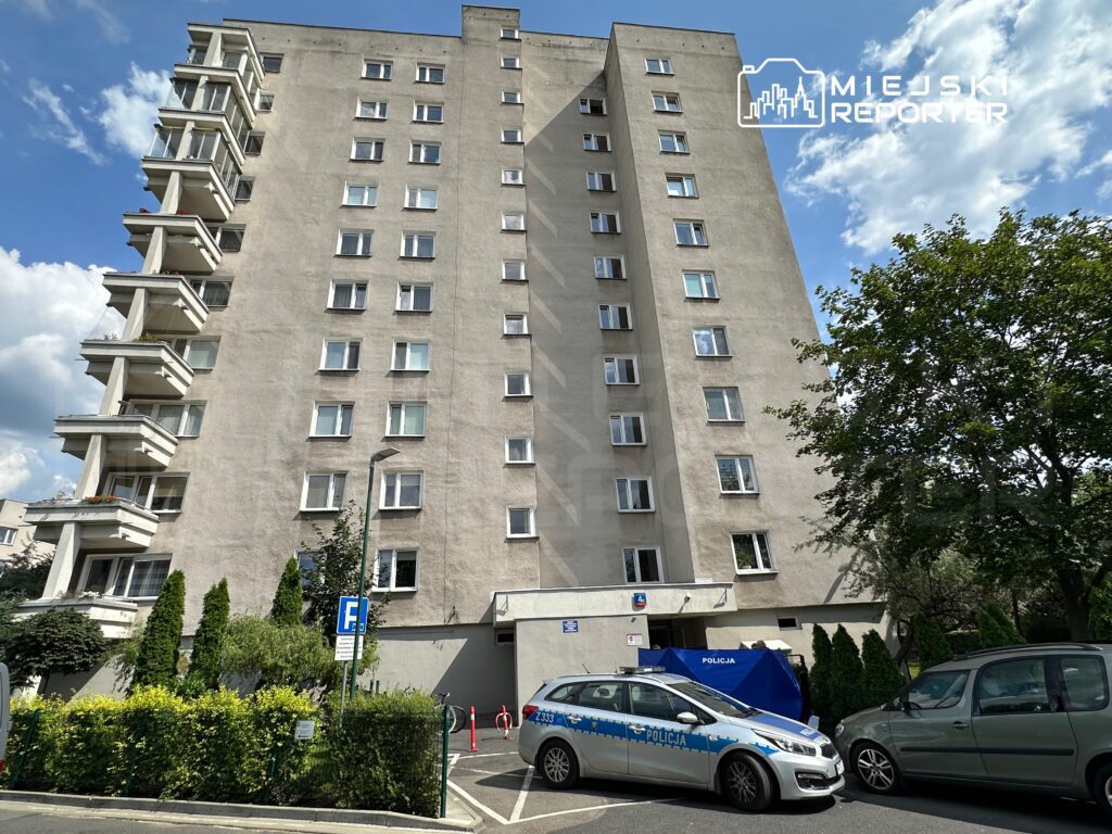 Wysoki, szary budynek mieszkalny z balkonami, w tle niebo z chmurami. Na parkingu policyjny samochód obok zielonych krzewów.