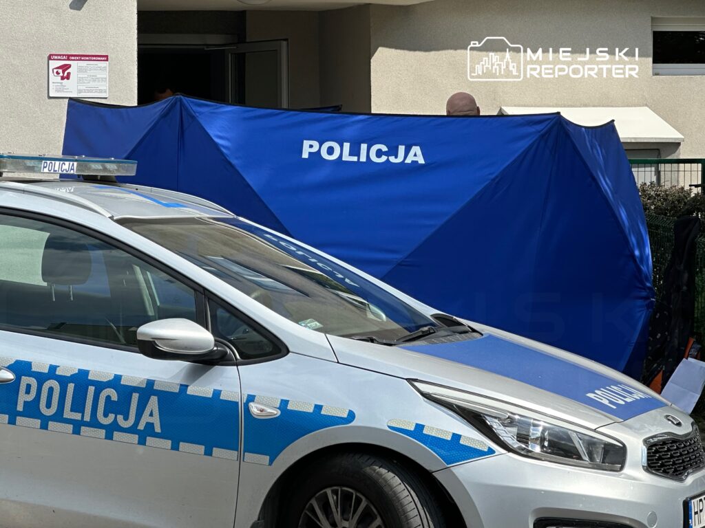 Policjant stojący za niebieską osłoną, obok radiowozu policyjnego, przy wejściu do budynku mieszkalnego.