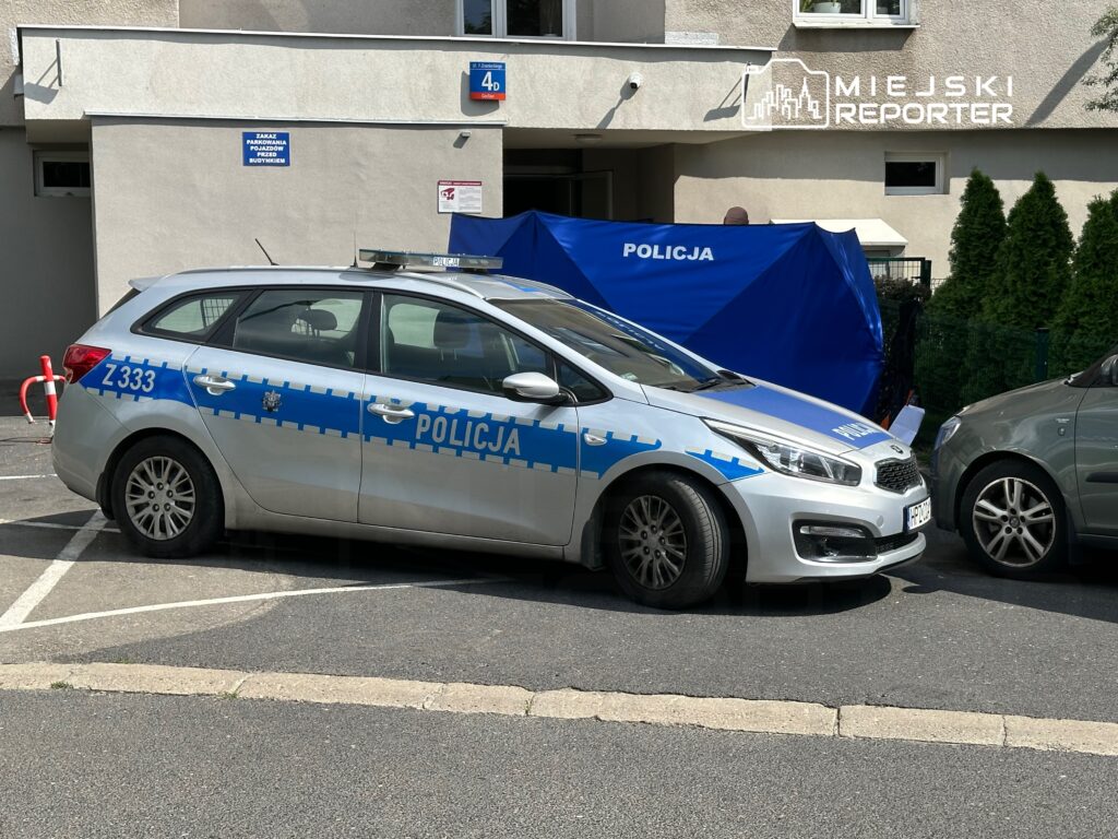 Policyjny radiowóz zaparkowany na osiedlowym parkingu obok niebieskiego namiotu z napisem "POLICJA".