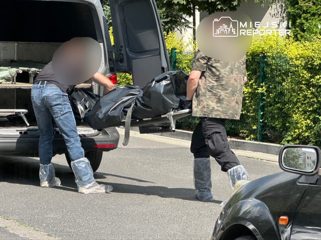 Dwaj mężczyźni w odzieży ochronnej przenoszą czarny worek z tyłu furgonetki na ulicy w otoczeniu zieleni.