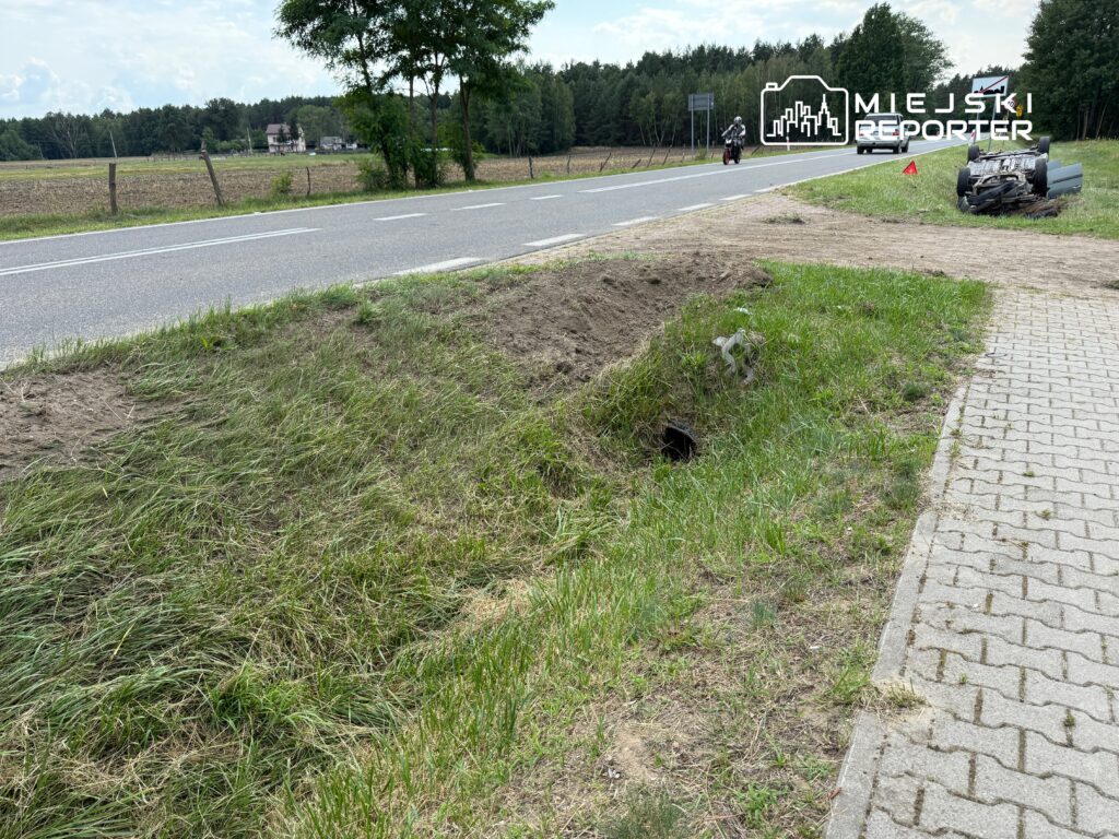 Na poboczu drogi leży przewrócony samochód, obok znajduje się motocykl, a w tle widać pola i las.
