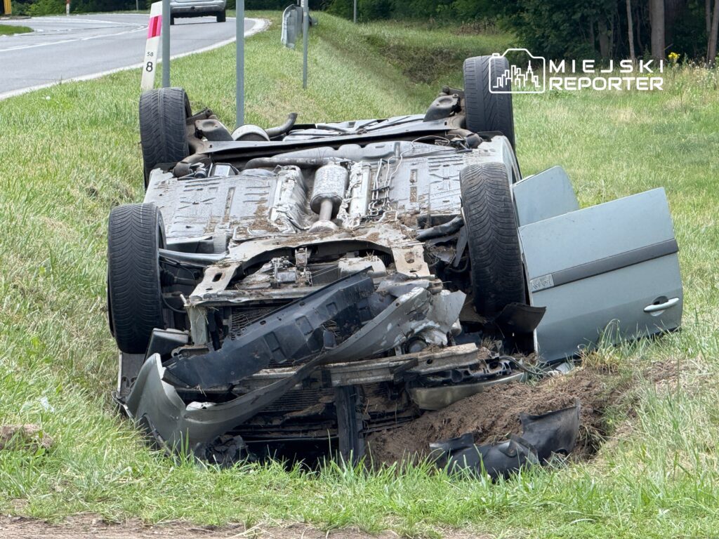 Samochód leżący na dachu w rowie obok drogi, z widocznymi uszkodzeniami i otwartymi drzwiami.