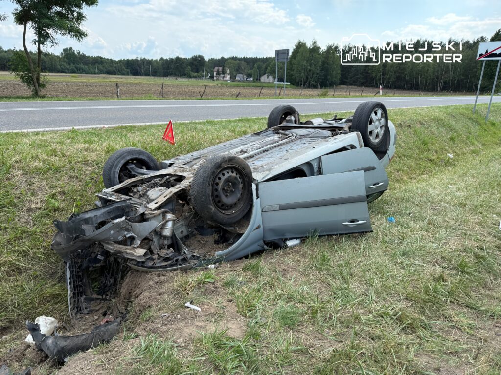 Przewrócone auto leży na boku w rowie przy drodze, obok znajduje się trójkąt ostrzegawczy.