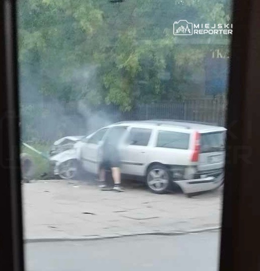 Osoba w krótkich spodniach bada uszkodzone, szare auto po wypadku, które uderzyło w przeszkodę na poboczu.
