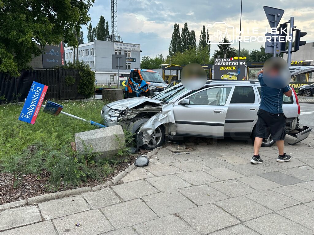 Zniszczony samochód osobowy stoi na chodniku obok przewróconego znaku drogowym, a mężczyzna sprawdza uszkodzenia.
