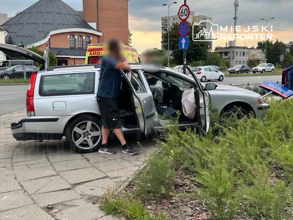 Mężczyzna w krótkich spodniach stoi przy otwartych drzwiach samochodu osobowego, obok znaku drogowym.