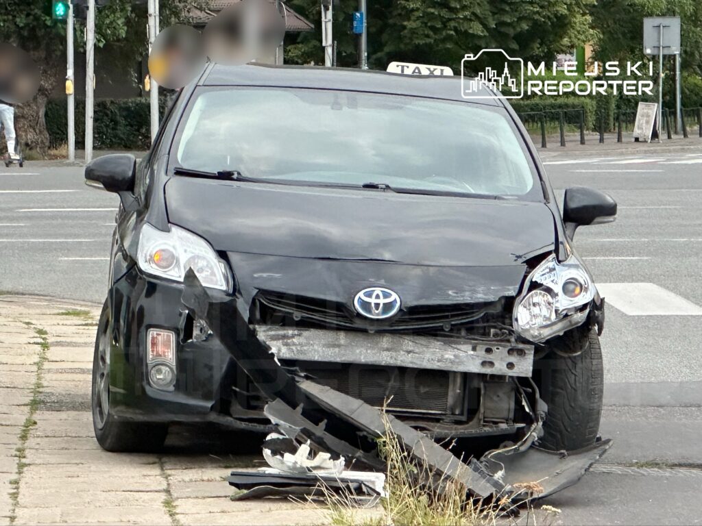 Czarny samochód osobowy marki Toyota z uszkodzoną przednią częścią stoi na chodniku w miejskim otoczeniu.