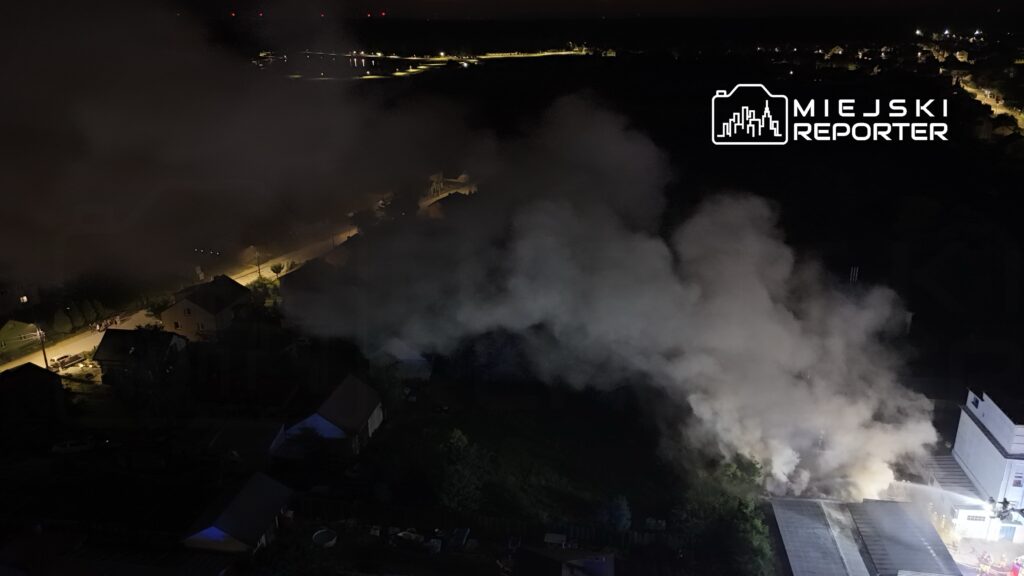 Gęsty dym unoszący się nad budynkiem w nocy, w tle oświetlona ulica z samochodami i domami.