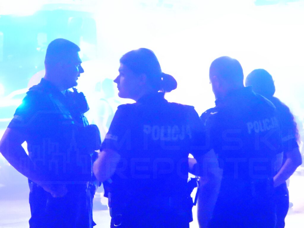 Czterech policjantów w mundurach rozmawia w zamglonym otoczeniu, z niebieskim światłem w tle.
