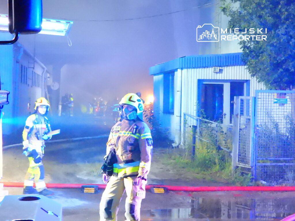 Strażacy w hełmach i maskach, z wężami gaśniczymi, pracują w zadymionym otoczeniu przy budynku.