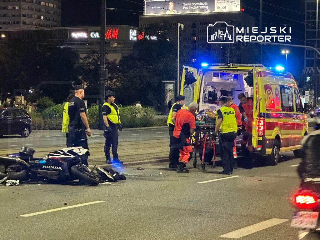 Na ulicy ratownicy medyczni transportują poszkodowanego na noszach do karetki, obok leży przewrócony motocykl.