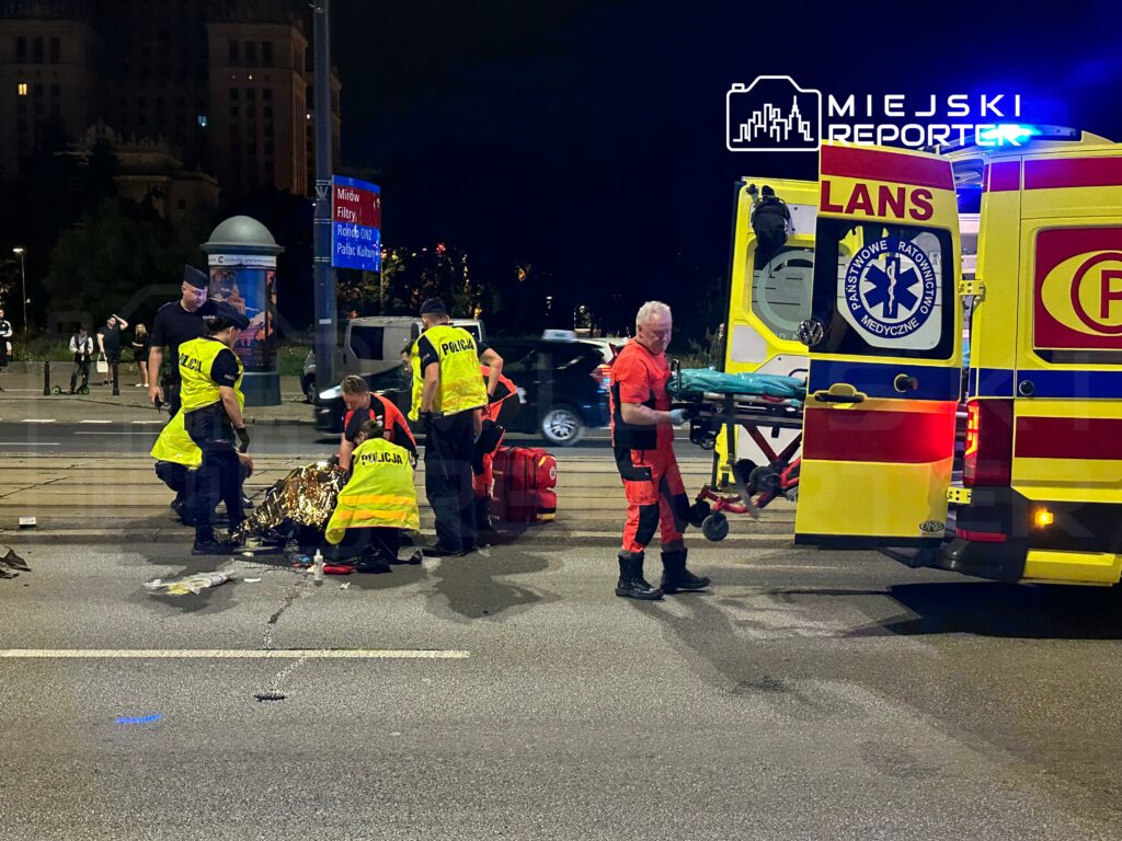 Grupa ratowników i policjantów udziela pomocy poszkodowanemu na ulicy w nocy, obok karetki.