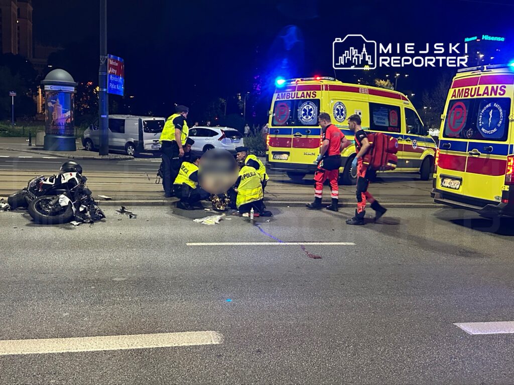 Motocykl leży na jezdni, a ratownicy medyczni udzielają pomocy poszkodowanemu w pobliżu ambulansów i policji.