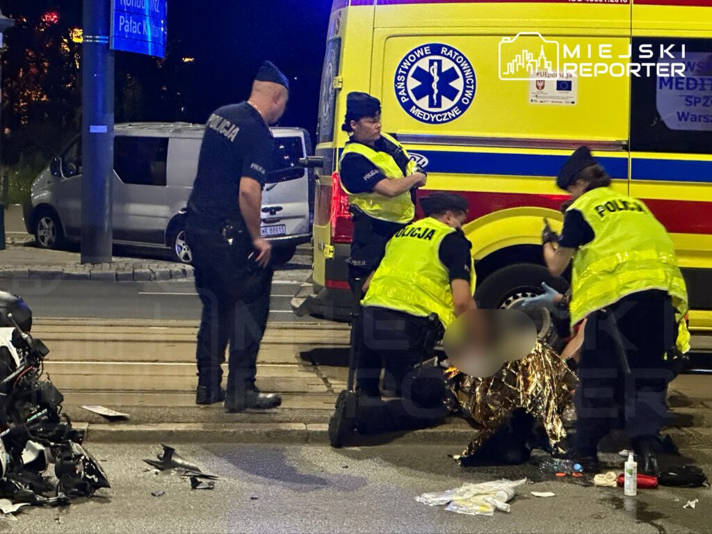 Grupa policjantów oraz ratowników medycznych udziela pomocy poszkodowanemu na ulicy, obok uszkodzonego motocykla.
