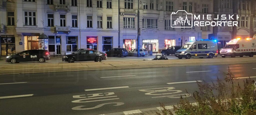 Na ulicy stoją samochody, w tym pojazd Bolt, obok policyjny van i karetka, w tle widać oświetlone witryny sklepowe.
