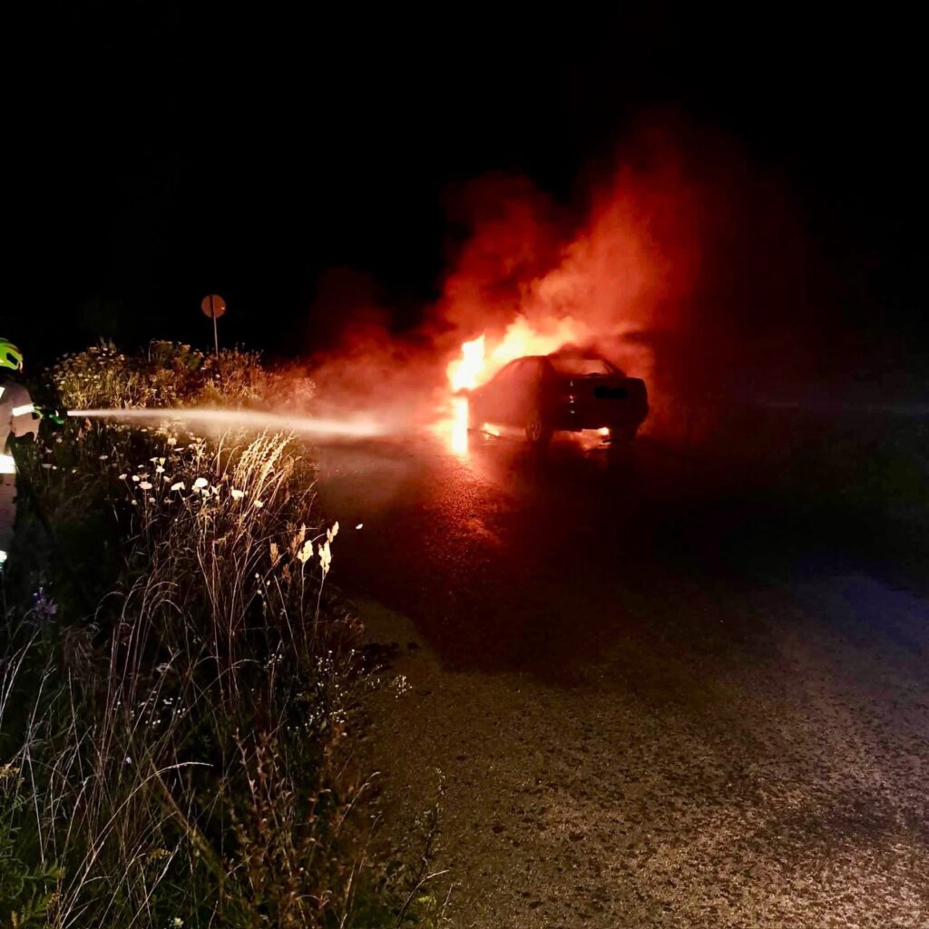 Strażak polewa wodą płonący samochód na drodze w nocy, otoczony dymem i wysoką trawą.