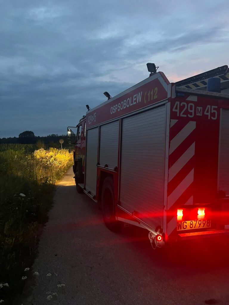 Czerwony wóz strażacki OSP Sobolew zaparkowany na wąskiej drodze, oświetlający otoczenie w świetle zachodzącego słońca.