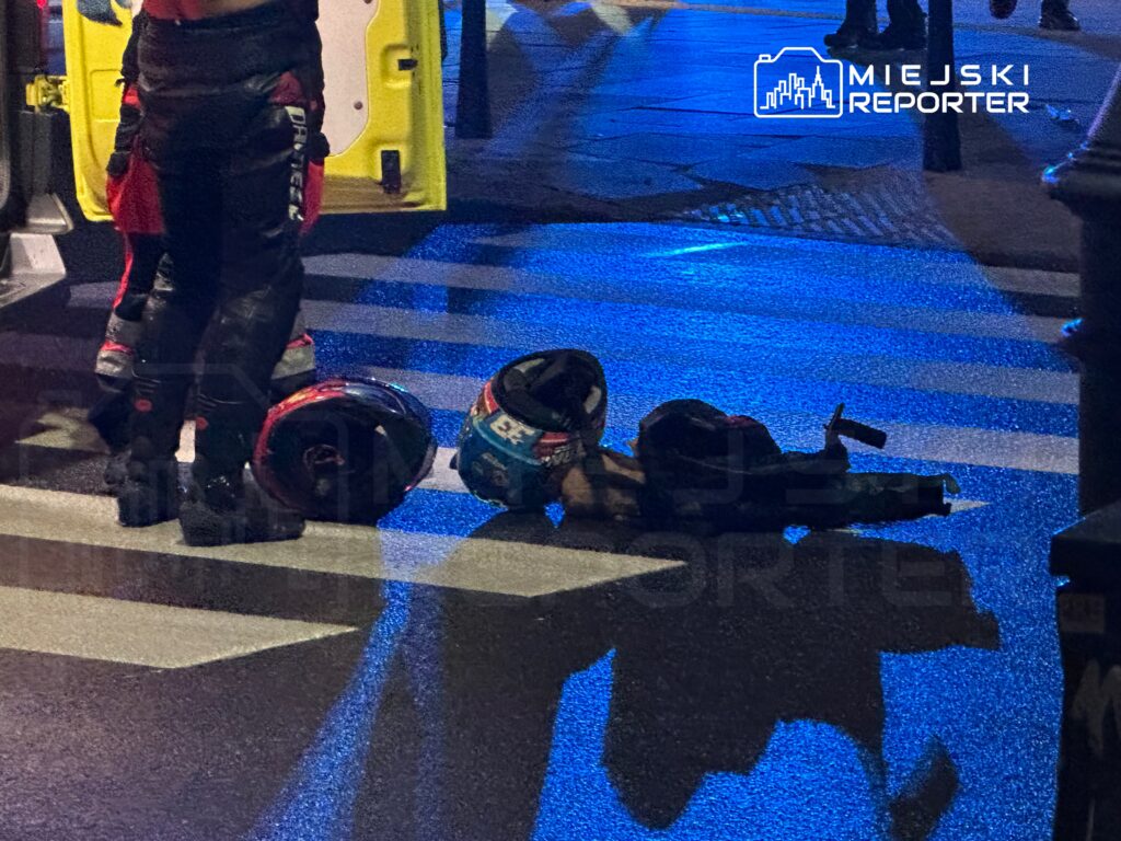 Motocyklista leży na jezdni obok dwóch kasków, podczas gdy ratownicy medyczni przygotowują się do interwencji.